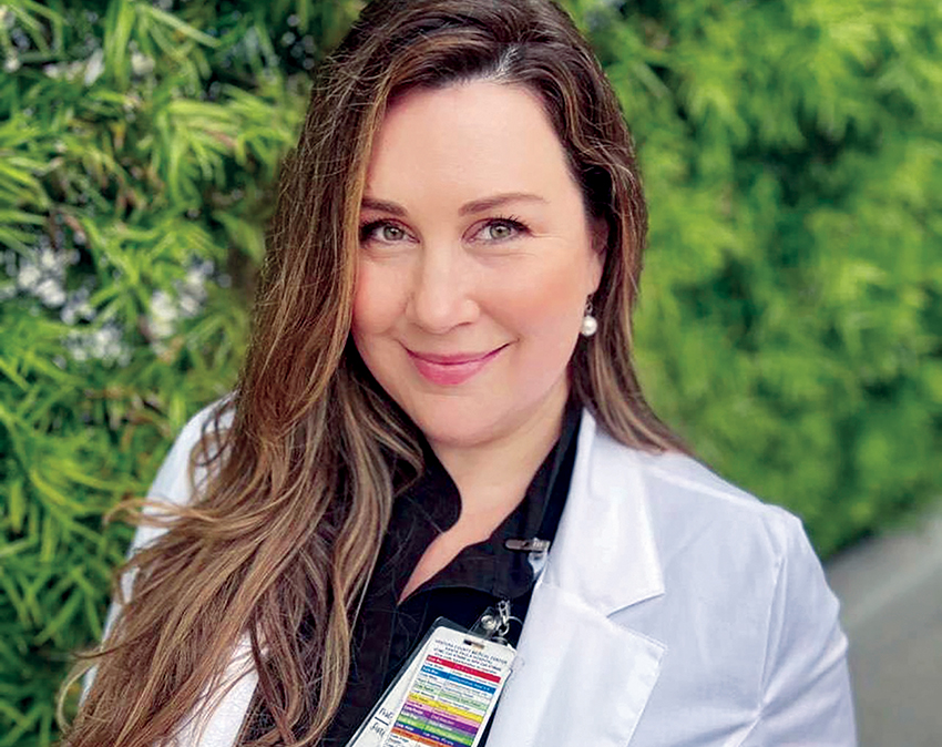 Smiling nurse in white coat stands in front of leafy background.