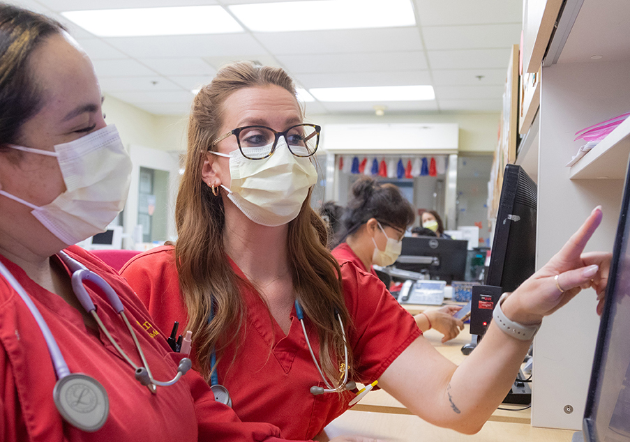 Nurse in red scrubs educates another nurse as she points to a computer screen