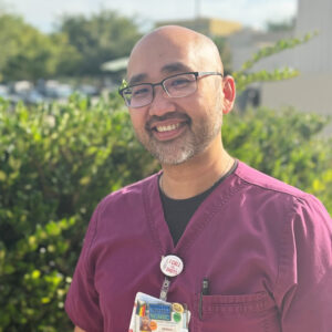 Male nurse in purple scrubs posting outside.