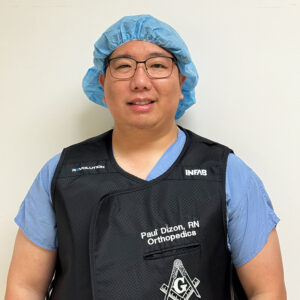 Male nurse wearing scrubs and a surgical cap standing against a beige wall.