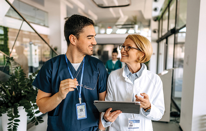 Male nurse in blue scrubs talking with a female doctor in white coat, walking down hospital hall.